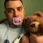 A grown man sits on the floor, saucking a baby nipple while playing with a teddy toy.