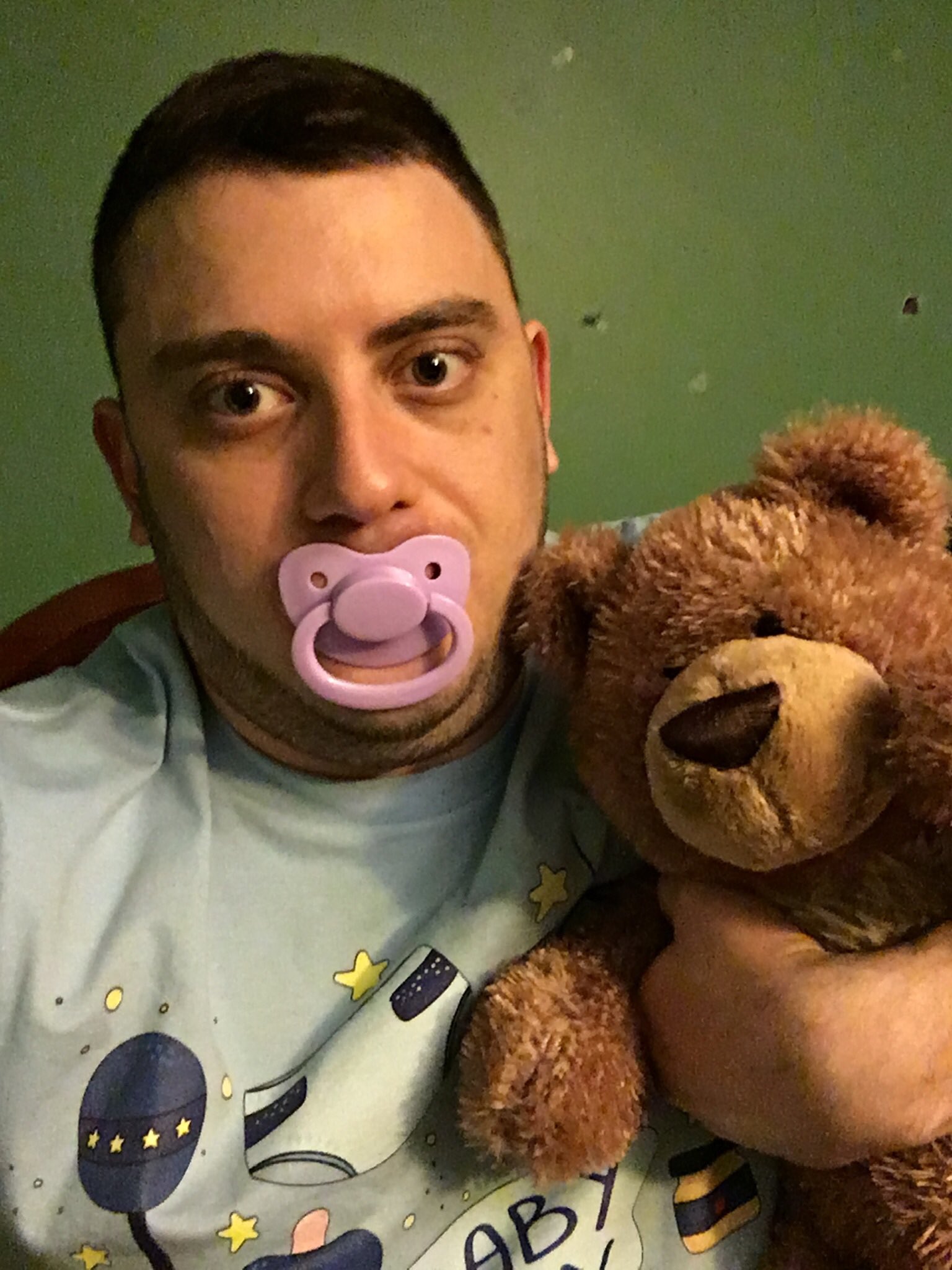 A grown man sits on the floor, saucking a baby nipple while playing with a teddy toy.