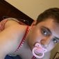 A baby boy lies in bed, sucking on a baby nipple while wearing a white diaper.