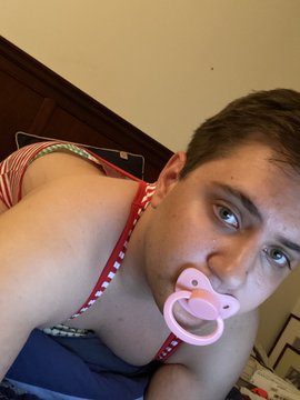 A baby boy lies in bed, sucking on a baby nipple while wearing a white diaper.