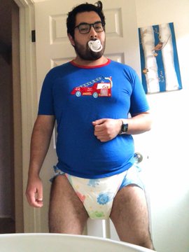 A grown man stands on the floor, saucking a baby nipple while dressed in a white diaper.