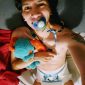 A baby boy is lying in bed, saucking a baby nipple while wearing a white diaper.